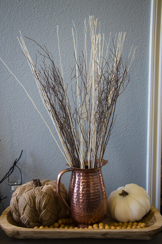 a hammered copper pitcher with dried branches, surrounded by a rope pumpkin, a white pumpkin and a wooden bead garland - all on bread board