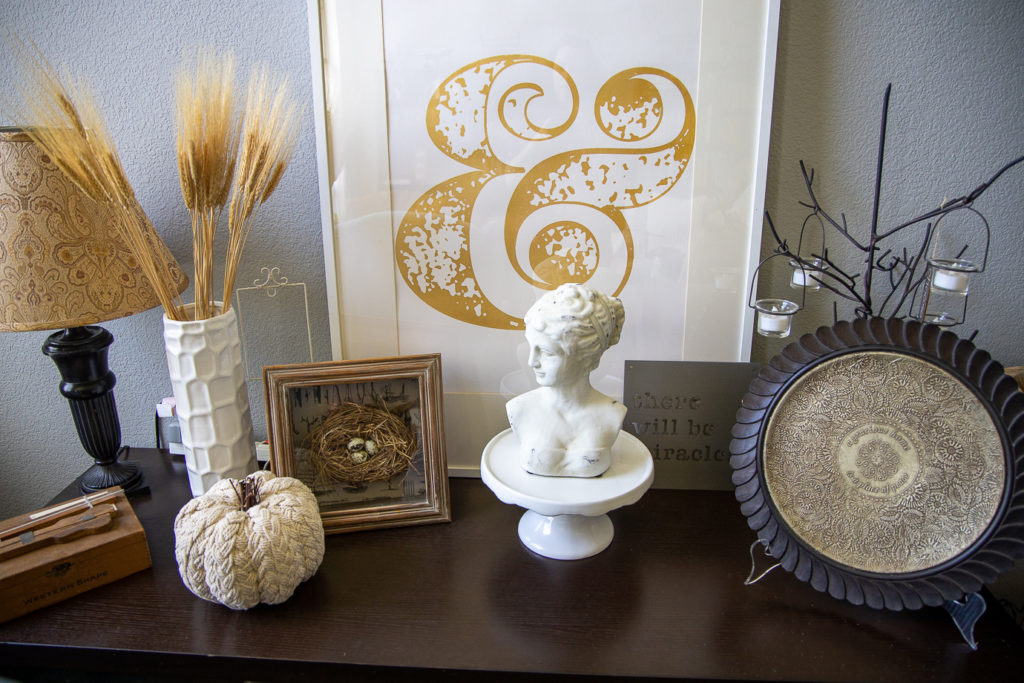 a group of items on a side table: a lamp, neutral rope pumpkin, a white vase with wheat, a shadow box with a bird's nest, and a white bust of a woman on a cake plate